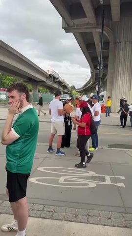 A group of people are gathered on a sidewalk next to a raised overpass or bridge. A man in a green shirt and black shorts is in the foreground, facing away from the camera and looking to his left. To his right, a group of people are interacting. A woman in a red shirt and black pants is holding a phone and looking down at it. A man in a white t-shirt and khaki shorts is standing next to her, holding a large stuffed animal that appears to be brown and fluffy, with green leafy material attached to its head. Several other men in white t-shirts are also present. One man is wearing a white baseball cap. Another man is wearing a blue baseball cap. The ground is paved, and a bicycle symbol is visible on the pavement. Beyond the people, there are vehicles and more of the overpass structure. The sky is cloudy.