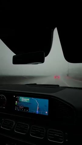 The video shows the interior of a car driving in the rain on a highway. The windshield wipers are active, and the visibility outside is poor due to fog and rain. The car's dashboard is visible, with a navigation screen displaying a map. There are other cars on the road ahead, their taillights visible in the distance. The side mirror shows a glimpse of the rainy landscape. The overall mood is one of travel in inclement weather.