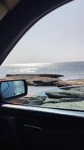 The video shows a view from inside a car looking out a window at a rocky coastline. The sun is shining brightly, reflecting off the water. In the foreground, a car's side mirror is visible, showing a reflection of the car's interior and the road. The ocean is in the background, with a clear, light-colored sky above it. There are several large rocks and boulders along the shoreline, with small pools of water between them. The overall scene is tranquil and scenic.