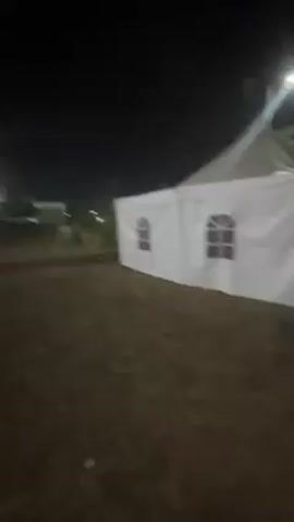 The video shows an outdoor setting at night, with a brick-paved area. A large white tent is visible on the right side of the frame. In the distance, there is a building illuminated by lights. People are walking around, and there appears to be some activity in the background, with vehicles visible. The camera moves, showing different angles of the area. The overall lighting is dim, with streetlights providing the primary illumination.