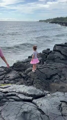 A young girl wearing a pink tutu stands on jagged black volcanic rock near the ocean. Waves crash against the shore, and lush green trees line the coastline in the distance. A person's hand, wearing a pink shirt, reaches into the frame from the left, seemingly interacting with the girl. The sky is cloudy but bright.