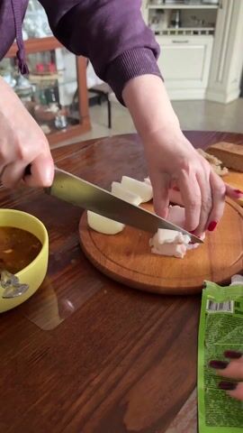 A person is preparing a meal on a wooden table. They are chopping white fat and onions on a round wooden cutting board with a large silver knife. To the left of the cutting board is a yellow bowl containing a brown soup with visible pieces of vegetables and meat. A silver spoon rests inside the bowl. The person has red nail polish. The table is made of wood and has a clear plastic protector on part of its surface. Background details include pieces of bread, a tube of what appears to be a food product, and a china cabinet in the back.