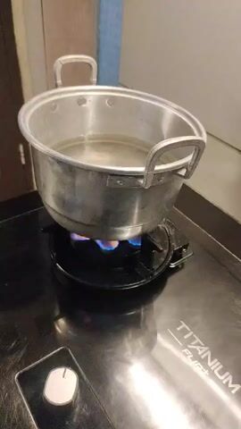 A medium shot captures a pot of water heating on a gas stove. The pot is made of aluminum and has two handles. The stove has a black glass top and a single knob on the front. The gas burner is lit, producing a blue flame. The water in the pot appears to be clear and still. The background is blurred, but suggests a kitchen environment with a wall and a cabinet. The lighting is warm and natural.