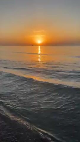 The video shows a serene sunset over the ocean. The sun is low on the horizon, casting a golden glow on the water. Gentle waves roll toward the shore, their surfaces reflecting the sunlight. There are a few boats visible in the distance, silhouetted against the bright sky. The beach in the foreground consists of dark-colored pebbles. The scene appears peaceful and tranquil.