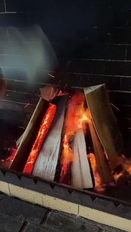 This video captures a close-up view of a roaring fireplace. The scene is set within a brick fireplace, with the dark brickwork forming the backdrop. A substantial pile of firewood is arranged in a teepee-like structure, with thick logs at the base and smaller pieces angled towards the center.

The heart of the scene is the intense fire burning within the logs. Flames are visible in vibrant shades of orange and red, licking at the wood and creating a warm, inviting glow. Sparks dance and fly from the burning embers, adding a dynamic element to the visual. Wisps of white smoke curl and drift upwards, obscuring parts of the fire and adding a sense of atmosphere.

The logs themselves appear to be seasoned, with visible grain and texture. Some of the wood shows signs of charring and glowing red from the heat. The foreground of the fireplace features a metallic grate, suggesting this is a functional fireplace designed for heating or cooking.

The lighting is dominated by the fire's glow, creating high contrast between the illuminated logs and the dark background. The overall impression is one of warmth, comfort, and the primal allure of an open flame. The setting appears to be indoors, possibly a home or a rustic establishment, given the presence of a fireplace. The time of day is not explicitly indicated, but the intimate and cozy nature of a fireplace suggests an evening or a cooler time of day. There are no people visible in the frame, focusing the attention entirely on the fire and the wood. There is no visible text in the video.