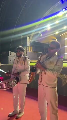 The video shows a band performing on a stage at night. There are three musicians visible. The person in the center is playing a saxophone and is wearing a white uniform with gold trim and sunglasses. To their left, another person in a similar uniform is playing a snare drum and also wears sunglasses. To the left of the drummer, a third person in the same uniform is playing a trumpet and wearing sunglasses. The stage has a red carpet and is lit with blue and white lights. In the background, there are large speakers and a stage set. The band appears to be part of an event or celebration.