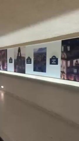 The video shows a person riding an escalator in what appears to be a subway or underground station. The escalator is long and has multiple sections. Other people are also riding the escalator in the same direction. The walls along the escalator are decorated with pictures of Paris, including the Eiffel Tower. The ceiling of the station is white and curved, with built-in lights. The overall lighting is somewhat dim.