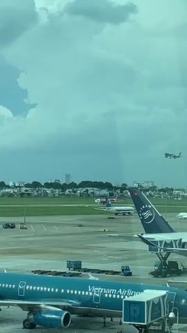 An aerial view of an airport tarmac. In the foreground, a blue Vietnam Airlines plane is parked at a gate, with a jet bridge attached. Further back, another plane with a blue and white tail fin is visible. In the middle ground, several other airplanes are on the tarmac, and a city skyline with buildings can be seen in the distance. In the upper right corner of the frame, a small airplane is in the process of landing or taking off against a backdrop of cloudy skies. The overall scene depicts a busy airport environment.