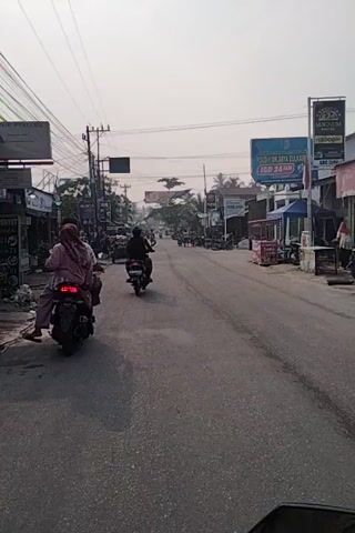 The video shows a street scene with several motorcycles and people. The camera is oriented vertically, giving the impression of a sideways view. In the foreground, a person on a red motorcycle is visible, facing away from the camera. Further down the street, other motorcycles and people are seen moving. Buildings line both sides of the street, with various signs and advertisements displayed. Power lines are also present overhead. The overall scene suggests a typical street in a populated area, possibly a market or a commercial district, with moderate activity. The lighting indicates it is daytime, likely with overcast skies.