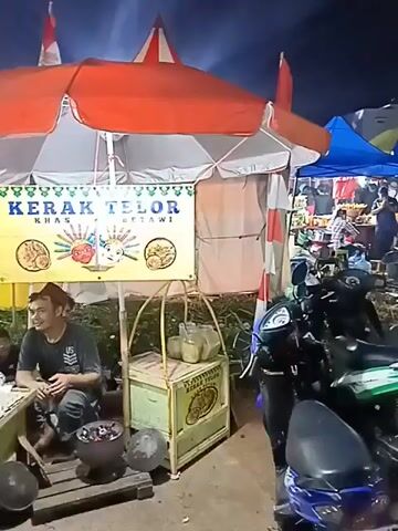 This is a video of a street food vendor in Indonesia, selling "Kerak Telor," a traditional Betawi dish. The vendor is a man wearing a dark t-shirt and a cap, sitting on a wooden pallet in front of his stall. The stall is set up under a large orange and white umbrella, with a yellow banner displaying the words "KERAK TELOR KHAS BETAWI." In front of the vendor, on a table, are several trays of eggs, a charcoal grill, and some cooking utensils. To the right of the stall, there is a parked motorcycle. In the background, other stalls and people can be seen, suggesting a busy marketplace or festival. The video is shot at night, with artificial lighting illuminating the scene.