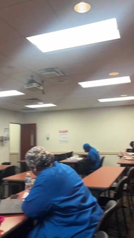 The video shows the interior of a classroom. The room contains several rows of desks and chairs, with two people seated at different desks, each wearing blue scrubs. The first person is working on a laptop with a pink bag on the desk, while the other is seated further back in the room. There is a whiteboard, a projection screen, and a podium at the front of the classroom. The ceiling has fluorescent lighting panels. The room appears to be sparsely occupied.