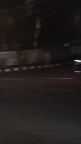The video shows a street scene at night. A motorcycle is seen traveling on the road. There are two people on the motorcycle, a driver and a passenger. The background shows a wall and some vegetation, possibly trees. There are streetlights visible, indicating it is nighttime. The overall impression is a passing view of a street with traffic at night.