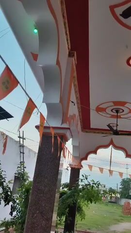 The video pans across the exterior of a building, likely a temple, adorned with orange pennant flags that read "Jai Shri Ram." The flags are strung along the eaves and hanging from architectural elements, creating a festive atmosphere. The building features white walls with orange trim and ornate decorative elements, including arched openings and a ceiling fan visible indoors. Green foliage from nearby plants is visible in the foreground, and a cloudy sky with hints of sunset can be seen in the background. A power line and a portion of a neighboring structure are also visible. The camera movement is smooth, suggesting a deliberate sweep of the area.