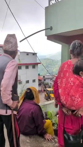 The video shows an outdoor scene from a slightly elevated perspective. In the foreground, three individuals are visible from behind. On the left, an elderly man wearing a cap and a vest over a pink shirt stands holding a cane. In the center, a person in a maroon shirt and yellow headscarf sits facing away from the camera. To the right, a woman in a bright pink outfit, with a child wrapped in a sling on her back, stands facing away. In the midground, a group of people are gathered in what appears to be a street or courtyard area. A yellow construction vehicle, possibly a backhoe loader, is positioned among them. Buildings of varying heights and architectural styles are visible in the background, with a hillside and trees beyond. The sky is overcast.