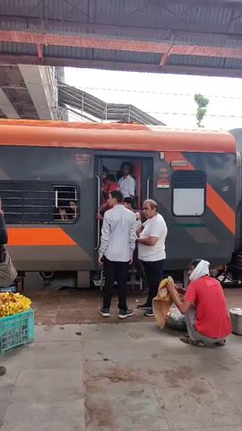 The video shows a train stopped at a platform. Several people are standing on the platform near the open door of a train car. One person is entering the train, while others are standing on the steps or just inside the doorway. On the platform, a man in a red shirt is sitting and appears to be preparing food or drink. Another man, wearing a light-colored shirt, is standing and talking to a man in a white t-shirt. A crate of bananas is visible on the left side of the frame. The train car is grey with orange and black accents. The platform appears to be made of concrete. The overall scene suggests a busy railway station.