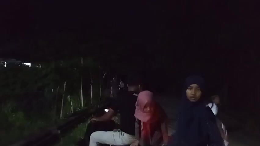 The video captures a group of people at night in what appears to be a rural setting in Geulanggang Baro, Indonesia. The lighting suggests it's nighttime, with artificial lights from a street lamp and motorcycle headlights illuminating parts of the scene.

The subjects are primarily people, with several individuals visible, some on motorcycles and others standing or walking. Two women are prominent in the foreground of the first frame, both wearing hijabs. One is in a dark blue hijab and the other in a pink hijab. A young boy in a black cap is also visible in the foreground of a later frame, looking directly at the camera. Other figures, some appearing to be children and adults, are scattered throughout the scene, many obscured by darkness or motion blur.

The activity appears to be a gathering or movement of people, possibly on their way somewhere or having just arrived. The presence of motorcycles suggests transportation is involved. The setting seems to be a road or pathway lined with trees and vegetation, with a building or structures visible in the background illuminated by artificial light.

Notable details include the time of day being night, the presence of several motorcycles, and the use of artificial lighting. The emotions are difficult to discern clearly due to the lighting and motion, but the boy in the foreground seems alert. No visible text is present in the video.
