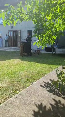 A person stands behind a podium in a grassy yard, addressing a small group of people gathered on the steps of a building. Sunlight filters through the leaves of a tree in the foreground, casting shadows on the ground. A speaker is positioned to the right of the podium.