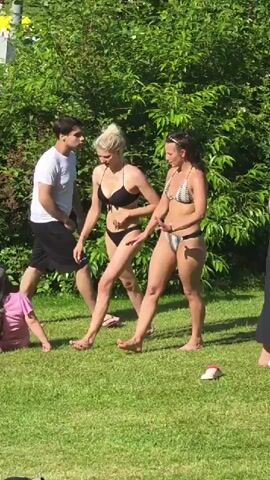 This media file captures a scene at an outdoor swimming pool area, likely in Lüdenscheid, Germany. The setting is a grassy area adjacent to a pool, with rows of lounge chairs and umbrellas visible in the background. Lush green bushes and trees frame the scene. The time of day appears to be daytime, with bright sunlight suggesting fair weather.

The primary subjects are people enjoying a day at the pool. In the foreground, two women in bikinis are walking across the grass. One has blonde hair styled in a bun and wears a black bikini. The other has darker hair and wears a patterned bikini. To their left, a child in a pink outfit is lying on the grass. Further back, various individuals are seen relaxing on lounge chairs, sunbathing, and socializing. A man in a white t-shirt and shorts is also visible walking.

The activity is typical for a public pool on a sunny day: leisure and recreation. People are sunbathing, resting, and likely preparing to swim or having just finished. The overall mood conveyed is relaxed and summery. There are no visible text elements within the media file.
