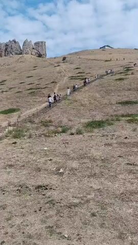 A group of people are walking up a dirt path on a dry, grassy hillside. There are large rock formations at the top of the hill. A silver car is parked at the bottom of the hill. The sky is blue with some white clouds.