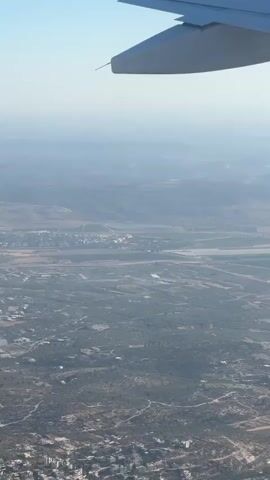 The video shows an aerial view from an airplane window. The top of the airplane wing is visible in the upper portion of the frame, extending across the top edge. The landscape below consists of rolling hills and a patchwork of agricultural fields, with some patches of trees and scattered buildings. A body of water, possibly a lake or reservoir, is visible in the distance. The overall atmosphere is hazy, suggesting a clear but somewhat distant view. The camera is static, and there are no discernible movements or actions occurring within the frame beyond the implied movement of the aircraft.