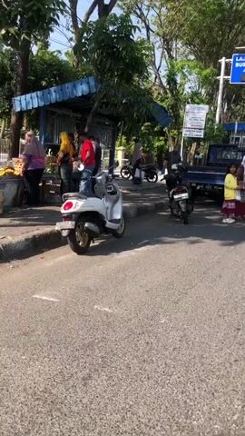 The video shows an outdoor scene on a paved road. Several people are present, some standing and some on vehicles. In the foreground, a white scooter is parked. To the right, a black pickup truck is stopped. Further back, a woman on a bicycle is moving away from the camera. There are also trees and a blue structure that appears to be a small building or kiosk. Signs are visible on poles and on the side of a building. The lighting suggests it is daytime.