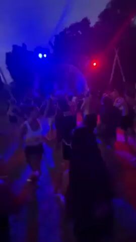 A group of people are dancing at an outdoor party at dusk. There is a DJ booth behind them with lights flashing blue and red. Behind the DJ booth are trees. People are standing around in the field, dancing and watching the lights.
