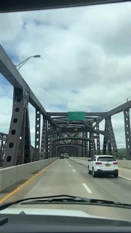 From inside a vehicle, a drive across a bridge with a metal truss superstructure is recorded. The bridge is painted dark gray. The road has a yellow line on the left and a white line on the right. There is a white car in the right lane of the road. There are a few other cars further ahead on the bridge. A green overhead sign states “The Hamilton Fish Bridge”. The weather is overcast with some blue sky visible. The video is stable.