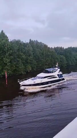 A luxury yacht with a white hull and navy blue accents cruises down a dark body of water, leaving a white wake. The yacht has multiple decks and windows. Along the left bank of the water is a dense line of green trees. The sky is overcast and grey. There is a red and white buoy in the water to the left of the yacht.