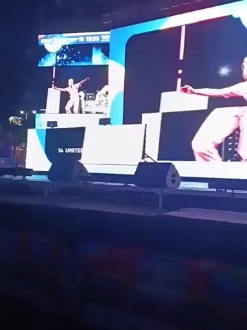 The video shows a stage with a large screen displaying a man in a pink jacket and white pants, who seems to be performing at a DJ console. The screen also shows an overlay of "14 UNITED KINGDOM." There are speakers in front of the screen on the stage. The setting is a concert or event, as indicated by the lighting and stage setup. The view is from a low angle, capturing the stage and the lower part of the audience or venue.