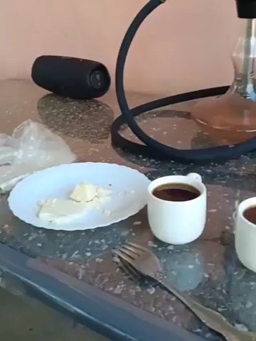 The video shows a table with a hookah, two cups of coffee, a plate of cheese, a salad of tomatoes and herbs, and a plastic bag. There is also a portable speaker on the table. The background consists of a wall and two potted plants. The camera pans across the scene, focusing on the various items on the table.
