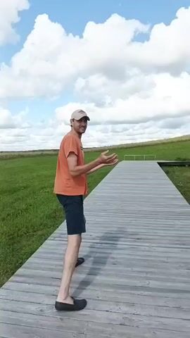 A man in an orange t-shirt and black shorts walks away from the camera on a wooden boardwalk. He is wearing a baseball cap and black slip-on shoes. The boardwalk is in a grassy field under a blue sky with white clouds. The man is smiling and looking over his right shoulder at the camera. The camera is positioned behind the man, following him as he walks down the boardwalk.