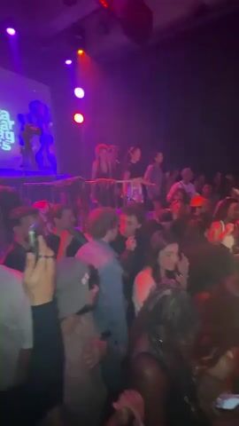A low-angle video captures a crowded dance floor with a DJ performing at a nightclub. The scene is lit with bright club lights, primarily pink and blue. A large screen displays visuals behind the DJ. The crowd is diverse, with people of various ethnicities. The DJ is positioned behind a setup with lit screens, presumably controlling the music. Many people in the crowd are moving and dancing, and some are holding up phones to record the performance. Above the dance floor is a balcony with additional patrons. The words "MISS YOU" are displayed in bright pink neon. Overall, the atmosphere is energetic and filled with motion.