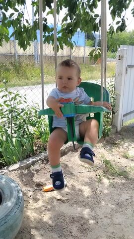 A young boy sits in a green swing in an outdoor yard. He is wearing a light blue and white striped shirt with a Bugs Bunny graphic, light blue shorts, and dark blue socks with green trim and a white Bugs Bunny graphic. He also has on dark blue shoes. The boy appears to be around two years old and has light brown hair. He is looking down and touching a green and blue toy attached to the swing.

The setting is a sunny outdoor area with sandy soil and patches of grass and weeds. A chain-link fence with some green foliage visible behind it runs across the background, along with a white metal fence. To the right of the swing, there is a toy that appears to be a plastic bottle with a white lid and an orange cap. A tire is visible in the bottom right corner of the frame. The sunlight suggests it is daytime, likely a warm day. The overall mood appears to be one of a child enjoying playtime outdoors.