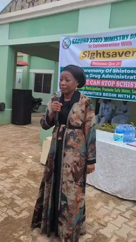 The video begins with a medium shot of a woman speaking into a microphone. She is wearing a headscarf and a long, patterned coat. Behind her, a green building with stone accents is visible. A banner with text is partially obscured but appears to be related to the Kaduna State Ministry of Health. To the left of the woman, a person in a green and yellow patterned outfit walks away from the camera. In the foreground, several tripods with cameras and microphones are set up, suggesting a press conference or media event. A man in a blue t-shirt and jeans is adjusting one of the cameras. Other individuals are seated in the background, some under a white tent. The ground is paved with interlocking bricks. The sky is overcast. The woman speaking is identified as Anita Buba.