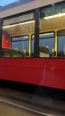 The video is taken from inside a car. The viewer is driving parallel to a tram. The tram is red and beige, and has the number 33 displayed. The tram has several windows, which are lit from the inside. It is raining, and there are droplets on the car window. The sky is overcast.