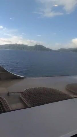 Here is a detailed description of the video:

The video presents a scenic view from a vessel or large ship, possibly a cruise ship. The perspective is from inside, looking out through a large window. The foreground shows parts of the ship's structure, including a white railing and metallic grates. 

Beyond the ship's elements, the primary focus is on the expansive view of a calm, blue sea. In the distance, there is a coastline featuring a series of green, mountainous islands under a partly cloudy sky. The sky is blue with puffy white clouds that add texture to the scene.

The camera remains steady, capturing the serene and somewhat hazy atmosphere. There are no identifiable people, but the perspective suggests a passenger or crew member is observing the scenery. Overall, the video presents a travel and nature scene.