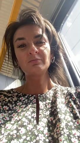 The video shows a woman in a floral dress recording a selfie video while on a train. She has dark hair and a nose ring. The train interior is visible, with gray seats and colorful seat cushions. Other passengers are sparsely seated throughout the train car.