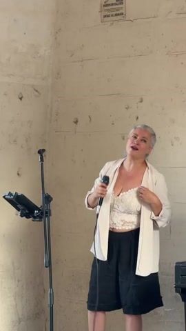 A woman with gray hair pulled back in a ponytail stands center frame, holding a microphone in her right hand and gesturing with her left. She is wearing a white lace crop top, a cream-colored blazer, and black shorts. She appears to be singing or speaking into the microphone, looking slightly upwards and to her left. To her left, a black microphone stand with a tablet attached is visible. The background is a textured, light brown wall with visible imperfections and some signage at the top right. The lighting is soft and natural.