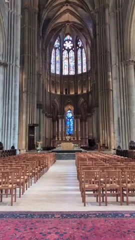 The video shows the interior of a large Gothic cathedral. Rows of wooden chairs are arranged on either side of a central aisle, leading towards an altar at the far end. The altar area is illuminated and features stained glass windows with vibrant colors, primarily blues and reds. The cathedral's architecture is characterized by tall, arched columns and intricate stonework. The lighting is dim, with spotlights illuminating specific architectural features and the stained glass. The overall impression is one of grandeur and solemnity.