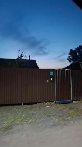 The video shows a blurry, exterior shot of a rural area at dusk or dawn. The sky is a deep blue with wispy clouds. In the foreground, a dark brown metal fence runs horizontally across the frame. Behind the fence, a dark, curved structure, possibly a roof or canopy, is visible. Trees and foliage are present on the right side of the frame. The camera is moving, creating motion blur throughout the video.