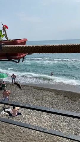 A person in a blue swimsuit stands in the ocean near a pebble beach. Waves crash on the shore. A red planter with a plant is visible in the upper left corner, attached to a thick rope that crosses the foreground. The sky is clear and blue. There are other people on the beach, some sitting and some standing.