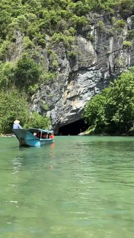 A boat is seen from behind moving on a river towards a large cave opening. The cave is dark and appears to be a natural rock formation. The boat is blue and has several people on it. The river is a bright green color, and the surrounding landscape is lush with greenery and trees. The sky is visible above, appearing to be a clear, bright day.